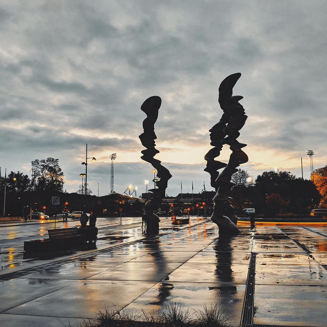 Two abstract stacked-figure sculptures reflected in a wet plaza surface under a dramatic cloudy sunset sky