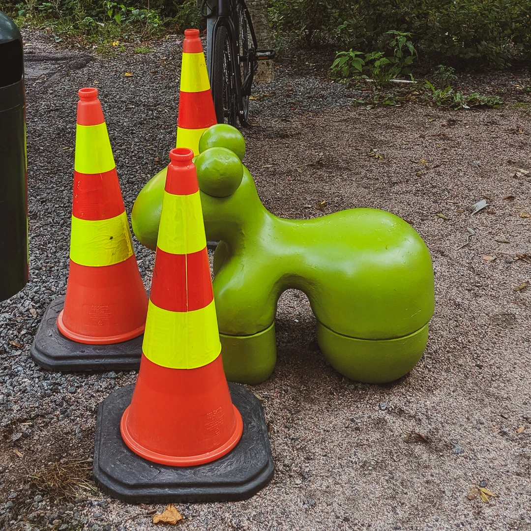 Two orange traffic cones standing next to a rounded green playground sculpture on a gravel surface